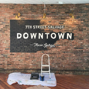 Mid process of installing the 7th Street Salvage Downtown mural. The white typography says 7th Street Salvage Downtown Macon, Georgia. The black background is halfway painted.