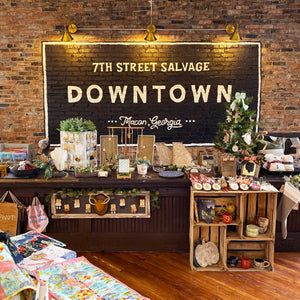 7th Street Salvage Downtown mural. a large black box with white typography inside say, "7th Street Salvage Downtown Macon, GA." In front of the mural is a general store counter with different vendor's products displayed on it for sale.