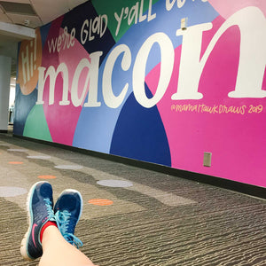 Ground view of a multicolored color block style painted mural with "Hi! we're glad y'all are in macon" hand lettered in white lettering with the word "macon" in larger, thicker font.