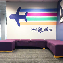 Load image into Gallery viewer, White wall with painting of a dark blue airplane with light blue, pink, green, and orange lines trailing behind it with text &quot;come fly with me&quot; in dark blue hand lettering
