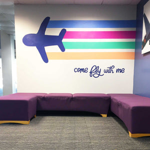 White wall with painting of a dark blue airplane with light blue, pink, green, and orange lines trailing behind it with text "come fly with me" in dark blue hand lettering
