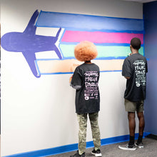 Load image into Gallery viewer, Two people paint green and orange lines under blue and pink lines trailing behind an airplane at the start of the macon airport mural
