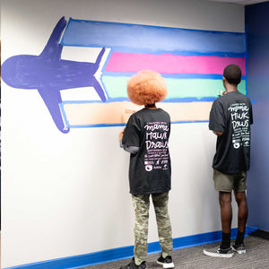 Two people paint green and orange lines under blue and pink lines trailing behind an airplane at the start of the macon airport mural