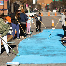 Load image into Gallery viewer, Adults and children holding paint rollers painting a section of road in blue paint
