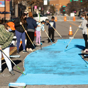 Adults and children holding paint rollers painting a section of road in blue paint