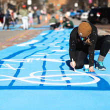 Load image into Gallery viewer, Woman kneeling over painted road painting a music note with white paint
