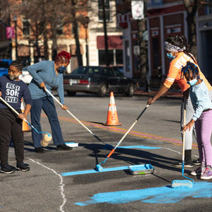 Two women and two children holding paint rollers and painting the sidewalk in blue paint