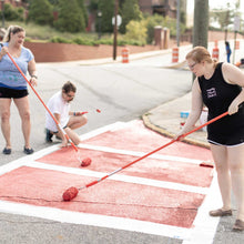 Load image into Gallery viewer, Three women painting red color onto the crosswalk. Two women are standing with paint rollers and one woman is kneeling over the painted area with paint roller in hand
