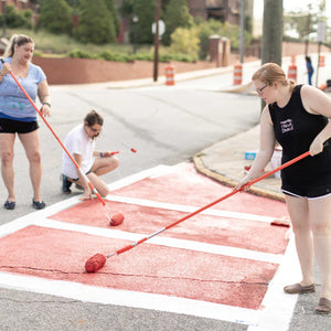 Three women painting red color onto the crosswalk. Two women are standing with paint rollers and one woman is kneeling over the painted area with paint roller in hand
