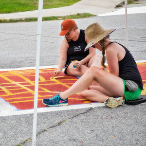 Two women sitting and painting red and orange street map design on crosswalk