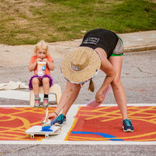 Load image into Gallery viewer, Photo of woman bent over crosswalk with paintbrush in hand. Little girl sitting on a stool in the background holding a large plastic cup sipping out of the straw watching with happy expression
