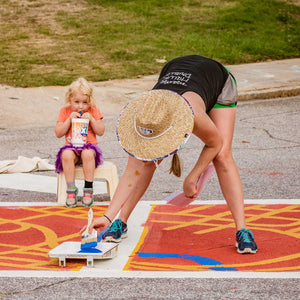 Photo of woman bent over crosswalk with paintbrush in hand. Little girl sitting on a stool in the background holding a large plastic cup sipping out of the straw watching with happy expression