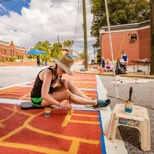Load image into Gallery viewer, Woman in hat sitting on a foam pad painting a red and orange street map design crosswalk
