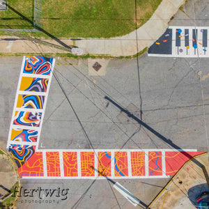 Aerial view photo of Mama Hawk's completed crosswalk mural, which is a map of Macon in burnt orang, golden rod yellow and a pop of blue for the Ocmulgee River. The photo also shows two of the other crosswalk murals by other artists.