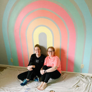 Erin from Mama Hawk Draws and her assistant Abby Noble wit in front of the completed rainbow for E's room. The rainbow is 3" stripes that gradate from buttery yellow, creamy orange, salmon pink, warm pink, soft green, and a sky blue. The wall is a dark gray behind the rainbow.