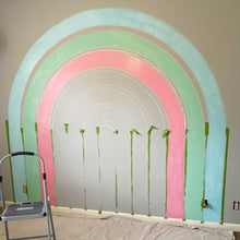 Load image into Gallery viewer, Half way through painting E&#39;s rainbow mural. The chalked out guidelines are still visible, but the first coat of sky blue, soft green, and warm pink are in place.

