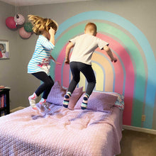 Load image into Gallery viewer, Two children jump on a bed in front of the completed rainbow in E&#39;s room. The rainbow is 3&quot; stripes that gradate from buttery yellow, creamy orange, salmon pink, warm pink, soft green, and a sky blue. The wall is a dark gray behind the rainbow.
