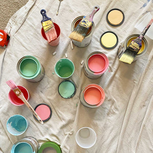 A look at the color pallet used for E's rainbow mural. You see open paint cans and cups filled with paint, plus paint brushes. Colors include soft green, warm pink, buttery yellow, salmon pink, and sky blue. 