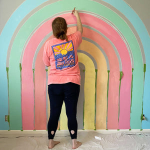 Abby works on the second coat for E's rainbow mural. She's painting the warm pink stripe and you can also see the first coats of the sky blue, soft green, salmon pink, creamy orange, and buttery yellow stripes. Between each stripe is a smaller stripe of the dark gray wall. 