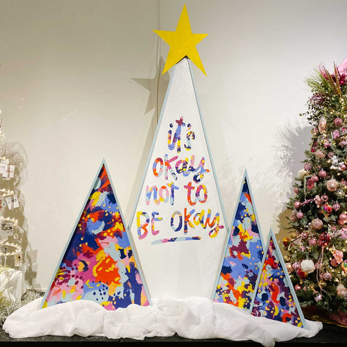Four brightly painted triangle trees sit in a museum on top of soft white fabric covered in glitter. The center tree is the largest and on top is a bright yellow star. On the tree is the phrase 