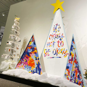 Four brightly painted triangle trees sit in a museum on top of soft white fabric covered in glitter. The center tree is the largest and on top is a bright yellow star. On the tree is the phrase "it's okay not to be okay" hand lettered in bright and vibrant colors. On the right flanks two smaller triangle trees, both brightly colored. On the left is one smaller, brightly colored triangle tree. Each tree is edged with a light blue wood that give the trees an extra dimension.