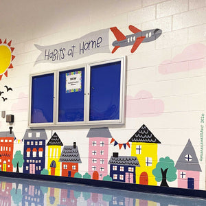 Ground view of "habits at home" mural with multicolored houses and buildings at the bottom of the wall and an airplane holding a banner with "habits at home" hand-lettered in black text.