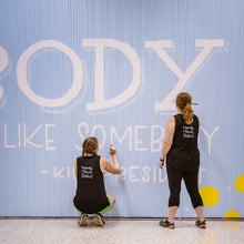 Load image into Gallery viewer, Close up of two women painting white letters on the Blue wall mural with white text that says &quot;be someone who makes everybody feel like somebody&quot;-kid president&quot;
