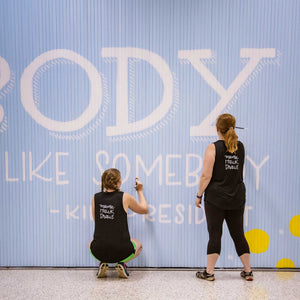 Close up of two women painting white letters on the Blue wall mural with white text that says "be someone who makes everybody feel like somebody"-kid president"