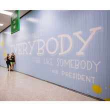 Load image into Gallery viewer, Full view of Blue wall mural with white text that says &quot;be someone who makes everybody feel like somebody&quot;-kid president&quot; with two women standing below the start
