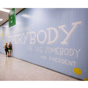 Full view of Blue wall mural with white text that says "be someone who makes everybody feel like somebody"-kid president" with two women standing below the start