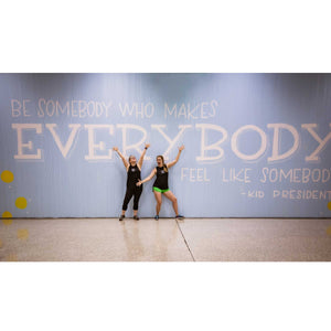 Two women posing in the center of  the Blue wall mural with white text that says "be someone who makes everybody feel like somebody"-kid president"