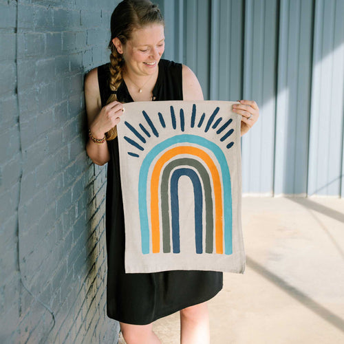 Erin from Mama Hawk Draws holds a 1.5' x 2' cream colored drop cloth with a painted rainbow on it. The rainbow has four stripes of the same width. From the inside out, the stripes are navy, dark gray, orange, and cyan blue. On the top of the rainbow are navy rays, radiating out at the top.