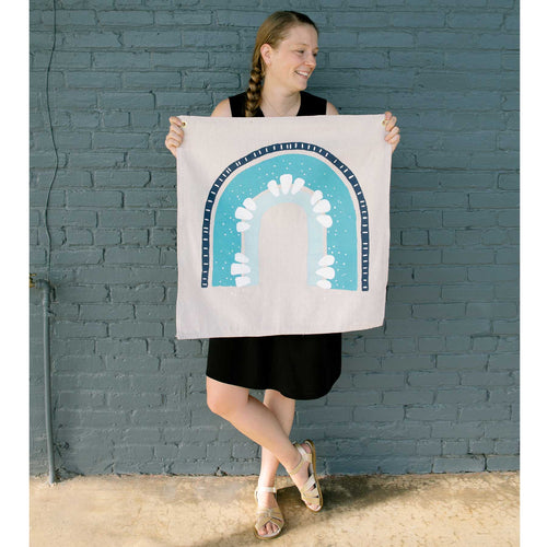 Erin from Mama Hawk Draws holds a 2'x2' cream colored drop cloth with a rainbow painted on it. The rainbow is different shades of blue, going from light blue to cyan to navy and has accents of white throughout the rainbow. Accents include dots and tick marks on the different rainbow stripes.