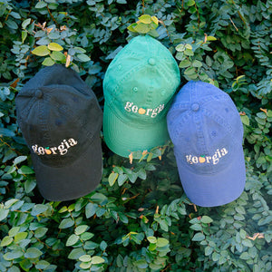 Three hats, black, green, and blue with "georgia" text with an orange peach as the "o" and greenery in the background