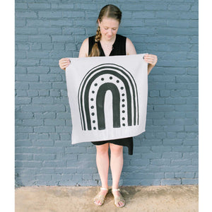 Erin from Mama Hawk Draws holds a 2'x2' cream colored drop cloth with a black rainbow painted on it. The rainbow has different widths of stripes. The interior stripe is the thickest, about 3", the next stripe is made of medium sized circles, the third stripe is about 2" thick, the 4th stripe is about 1" thick, and the last stripe is about 0.5" thick.