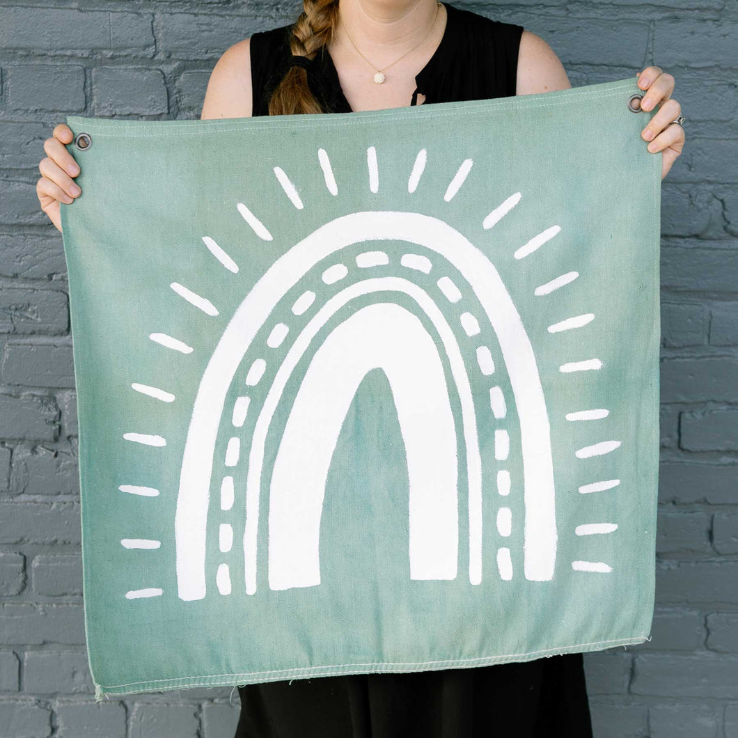 Erin from Mama Hawk Draws holds a 2'x2' green colored drop cloth with a white rainbow painted on it. The rainbow's four stripes have different thicknesses with one stripe made of dashes. Outside the stripes are rays around the rainbow.