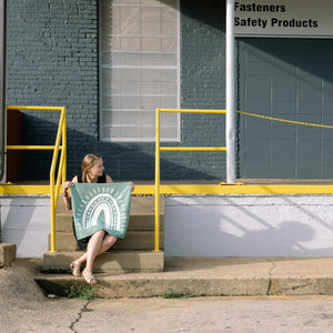 Erin from Mama Hawk Draws sits on a set of stairs outside an industrial area holding a 2'x2' green colored drop cloth with a white rainbow painted on it. The rainbow's four stripes have different thicknesses with one stripe made of dashes. Outside the stripes are rays around the rainbow.