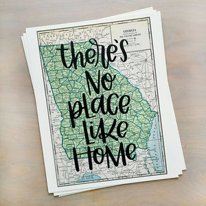 Stack of vertical papers on a table; top paper is a vintage style map design of the state of Georgia with black lettered text "there's no place like home" over the print.