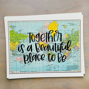 Stack of horizontal sheets of paper; paper on top is a blue, green, yellow, and pink flat map of the world with text "together is a beautiful place to be" in black lettering over the map print.