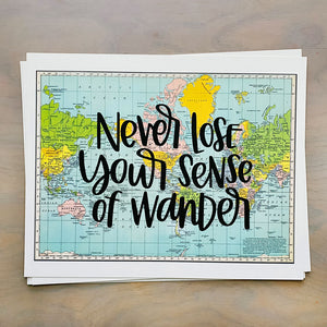 Stack of horizontal sheets of paper; paper on top is a blue, green, yellow, and pink flat map of the world with text "never lose your sense of wonder" in black lettering over the map print.
