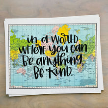 Load image into Gallery viewer, Stack of horizontal sheets of paper; paper on top is a blue, green, yellow, and pink flat map of the world with text &quot;in a world where you can be anything, be kind&quot; in black lettering over the map print.

