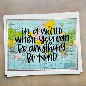 Stack of horizontal sheets of paper; paper on top is a blue, green, yellow, and pink flat map of the world with text "in a world where you can be anything, be kind" in black lettering over the map print.