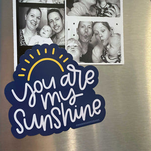 Photo of dark blue magnet with sun design with white hand lettering "you are my sunshine" holding up family photos on a refrigerator.