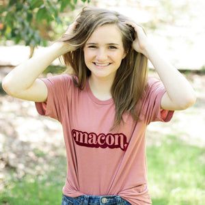 Photo of girl smiling wearing a pink tee with "macon" printed in the center with greenery in the background