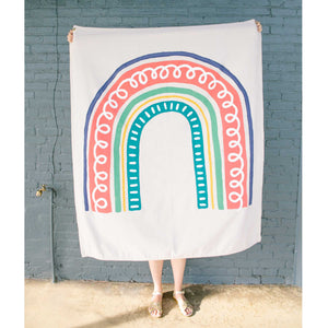 Erin from Mama Hawk Draws holds a 4'x5' cream colored drop cloth with a colorful rainbow painted on top. The rainbow has five stripes of varying color and thickness. From the inside, the first stripe is teal with white dashes accenting it. The second stripe is a thin yellow stripe. The third stripe is 2" green stripe. The fourth stripe is the thickest, about 5", in warm pink with a white squiggle through it. The last stripe is a blueish purple stripe.