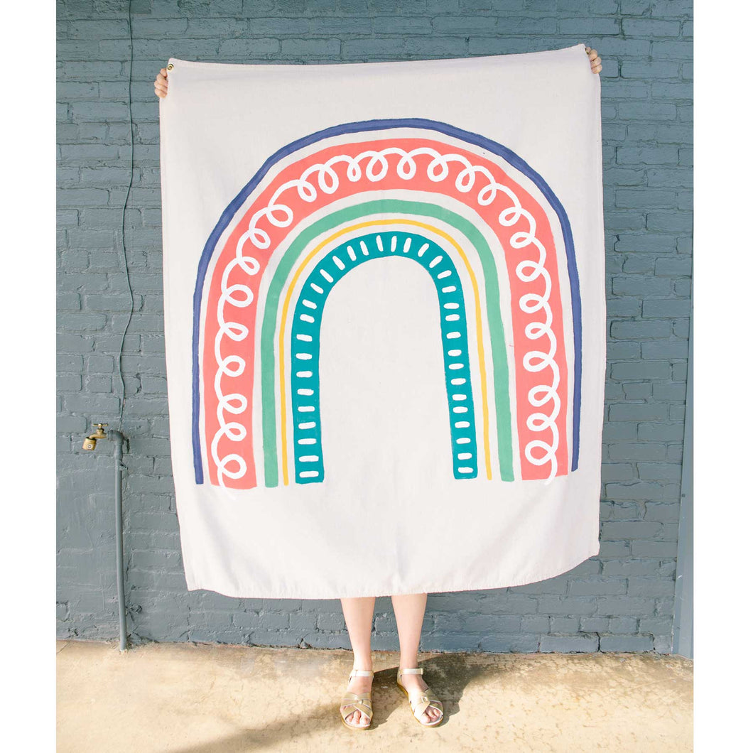 Erin from Mama Hawk Draws holds a 4'x5' cream colored drop cloth with a colorful rainbow painted on top. The rainbow has five stripes of varying color and thickness. From the inside, the first stripe is teal with white dashes accenting it. The second stripe is a thin yellow stripe. The third stripe is 2