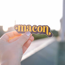 Load image into Gallery viewer, Up close photo of a white woman&#39;s hand holding a sticker over the Macon skyline. The sticker says &quot;Macon&quot; in orange in lower case serif font with ball flourishes on the &quot;m&quot; and the descender of the &quot;n.&quot; The word has a angled drop shadow in navy.
