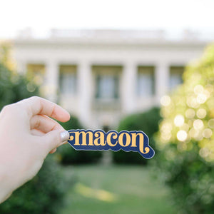 Up close photo of a white woman's hand holding a sticker in front of a blurred building. The sticker says "Macon" in orange in lower case serif font with ball flourishes on the "m" and the descender of the "n." The word has a angled drop shadow in navy.