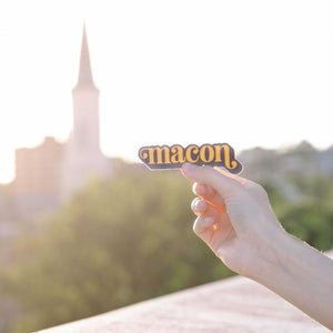 Up close photo of a white woman's hand holding a sticker over the Macon skyline. The sticker says "Macon" in orange in lower case serif font with ball flourishes on the "m" and the descender of the "n." The word has a angled drop shadow in navy.