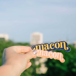 Up close photo of a white woman's hand holding two stickers over the Macon skyline. The stickers both say "Macon" in lower case serif font with ball flourishes on the "m" and the descender of the "n." The word has a angled drop shadow. The top sticker has orange lettering with a blue drop shadow. The bottom sticker has light pink typography and an orange drop shadow.